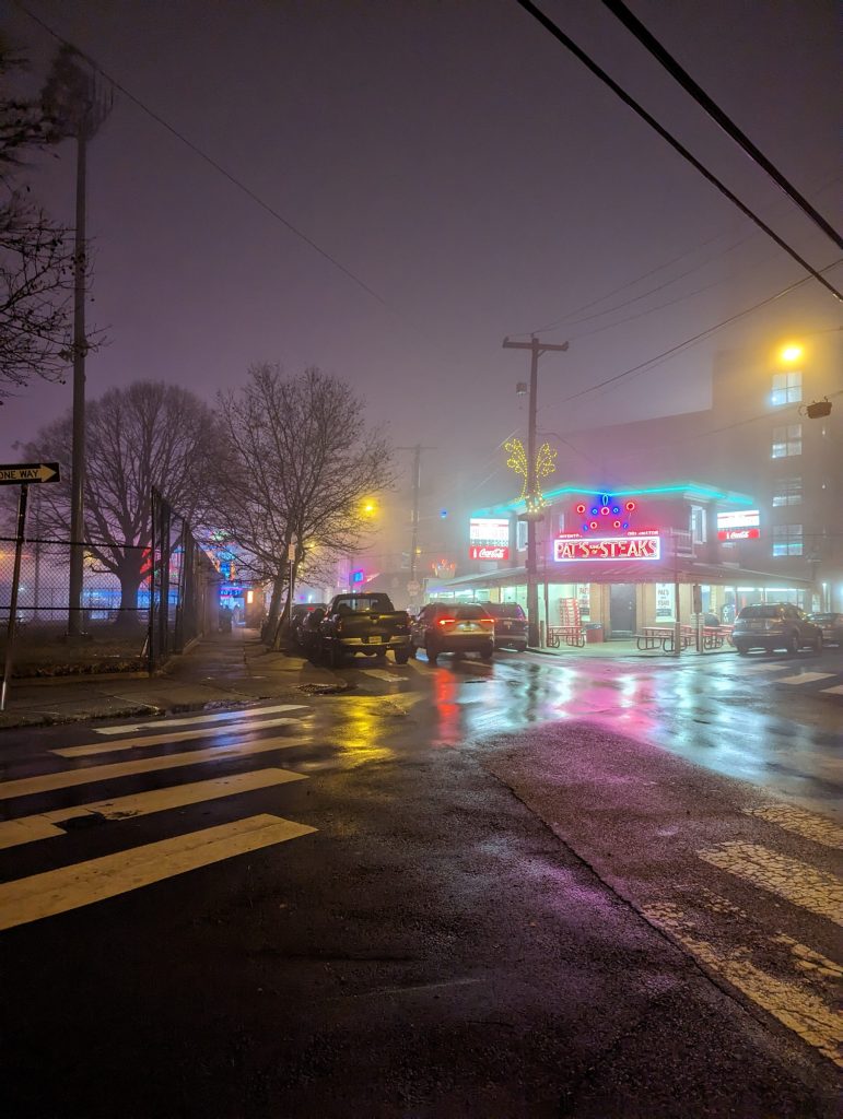 Streetlights and neon signs for Pat's Steaks in the foggy night, reflecting in the wet streets.
