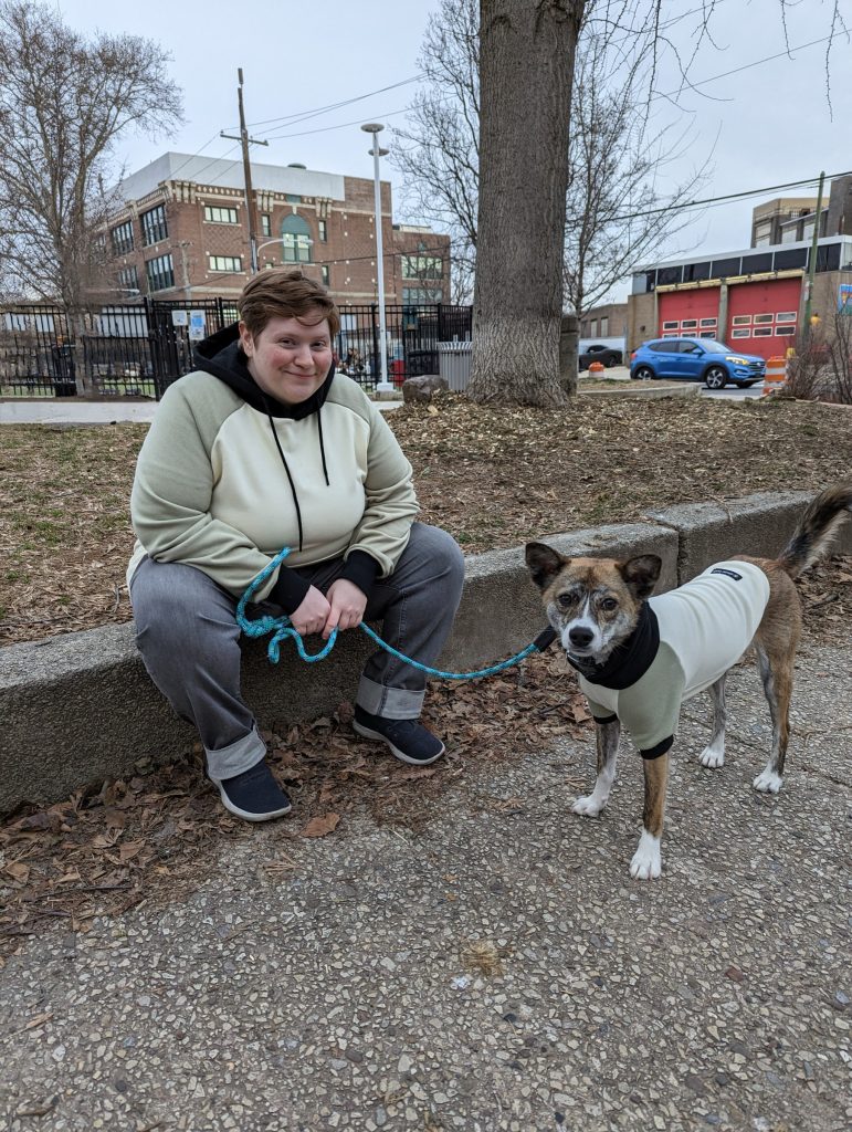 Mel, a redhead, and Noodle, a medium sized tan brindle dog, both in beige & green hoodies, sitting on the curb at the park.