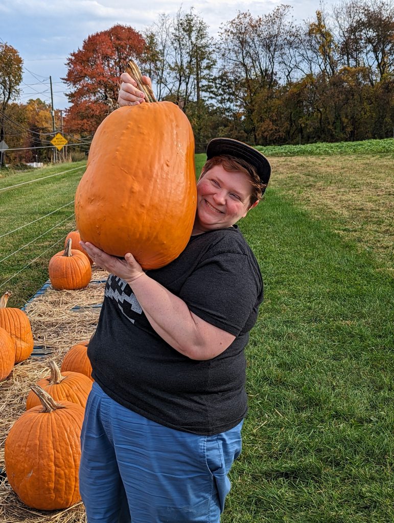 Mel holding up a very large pumpkin. The pumpkin is extra-tall, and easily 2-3 times the size of Mel's head.