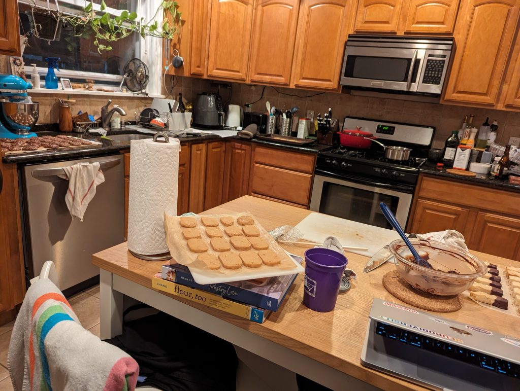 Our kitchen, with cookies on almost every surface. In the foreground, peanut butter cookies waiting to be baked and shortbreads dipped in chocolate. In the background on the counters are baked chocolate cookies and a tray of peanut butter cookies cooling.