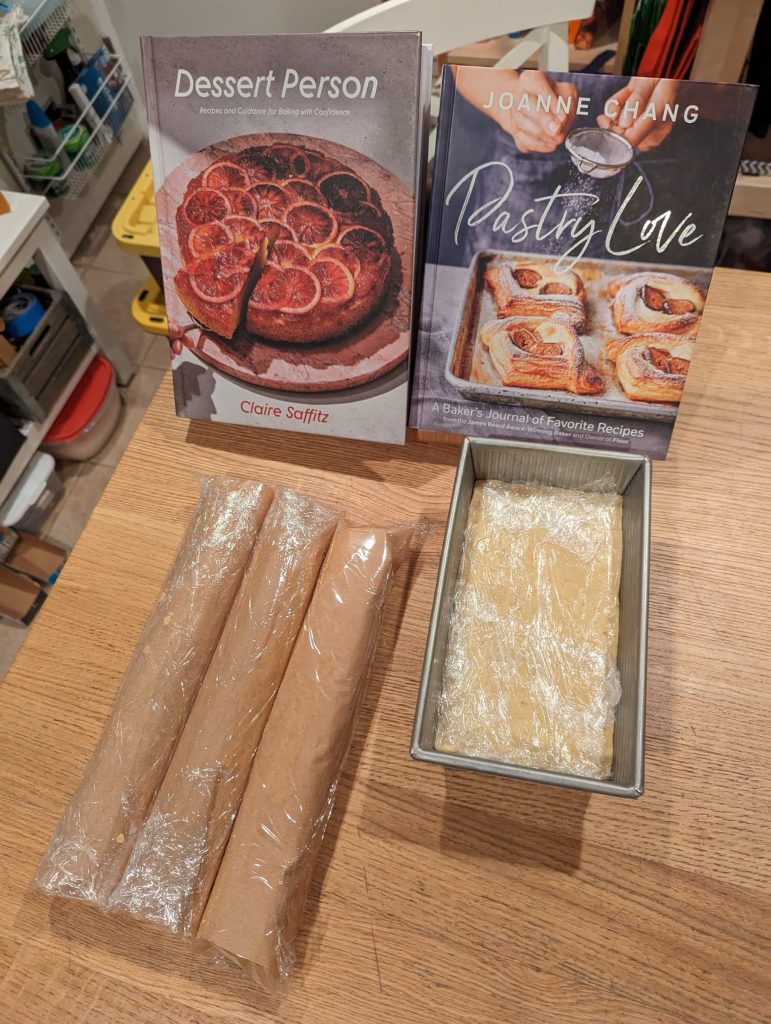 Two batches of dough. The first in 3 rolled tubes, in front of the Dessert Person cookbook, and the second pressed into a loaf pan in front of the Pastry Love cookbook.