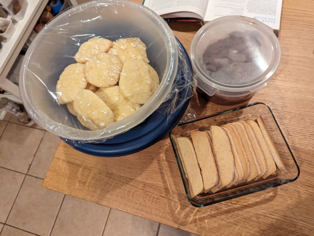 A bowl full of soft lemon cookies, a tray of rectangular lemon cookies, and a container of chocolate cookie dough.
