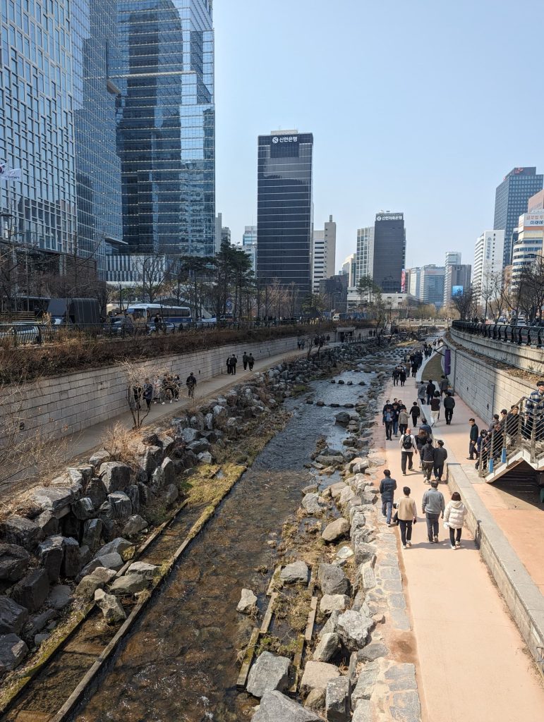 Looking down a stream in the middle of Seoul with pathways on both sides, full of people walking. There are retaining walls on each side, and beyond the walls, streets and skyscrapers.
