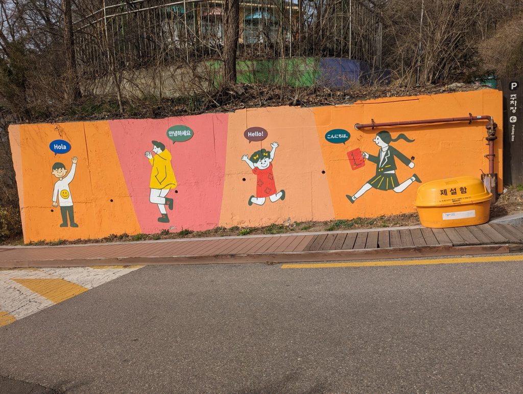 Colorful mural on a sidewalk wall, 4 people waving with "hello" in different languages.