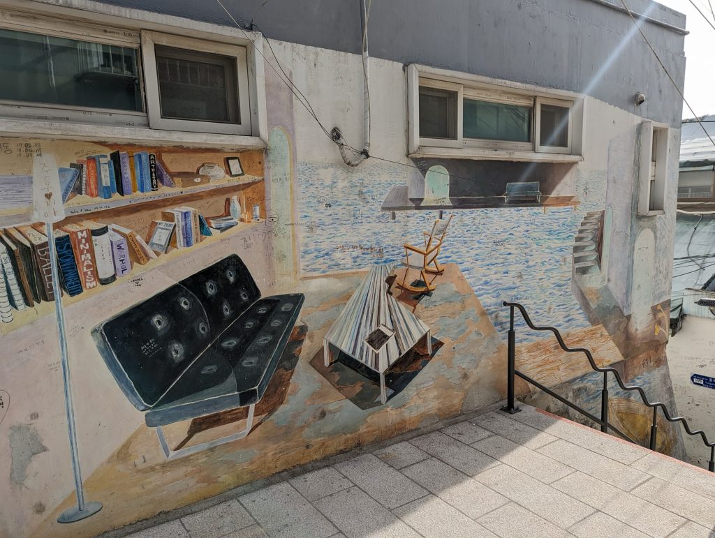 A mural on the side of a building next to the pathway's stairs. The mural incorporates the landing and stairs to look like an open living room overlooking water.