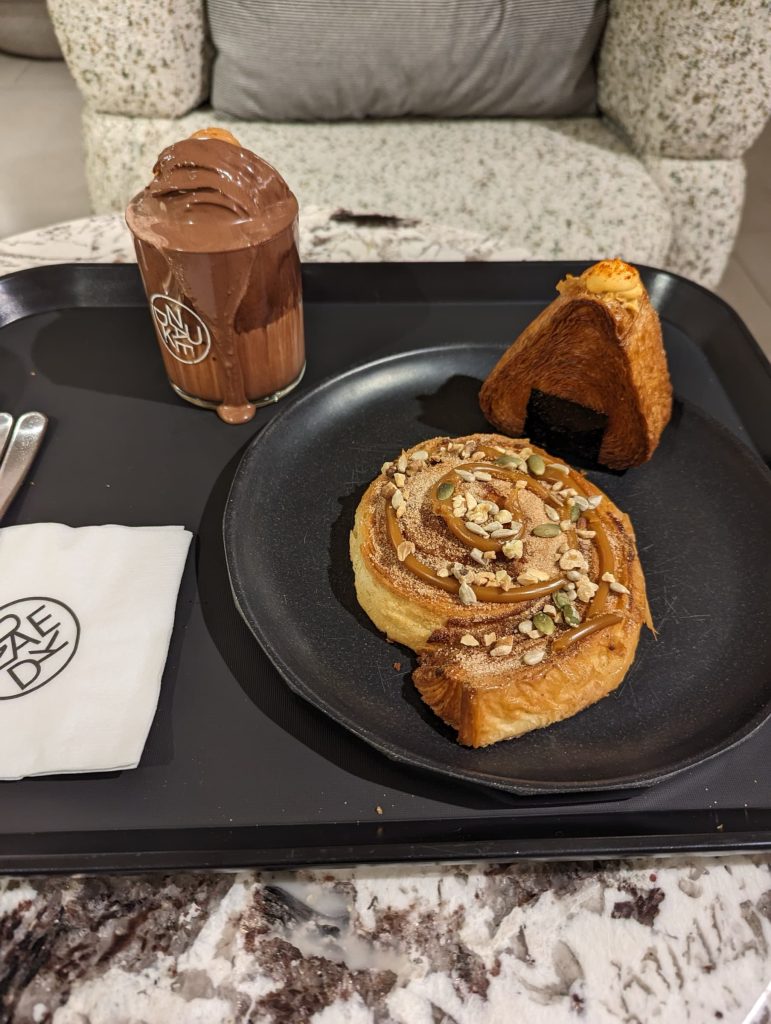 A tray with a chocolate drink, oniwassant, and spiral pastry. The drink is overflowing and topped with a croissant, coated in chocolate. The spiral pastry is topped with cinnamon, caramel, pepitas, and other nuts.
