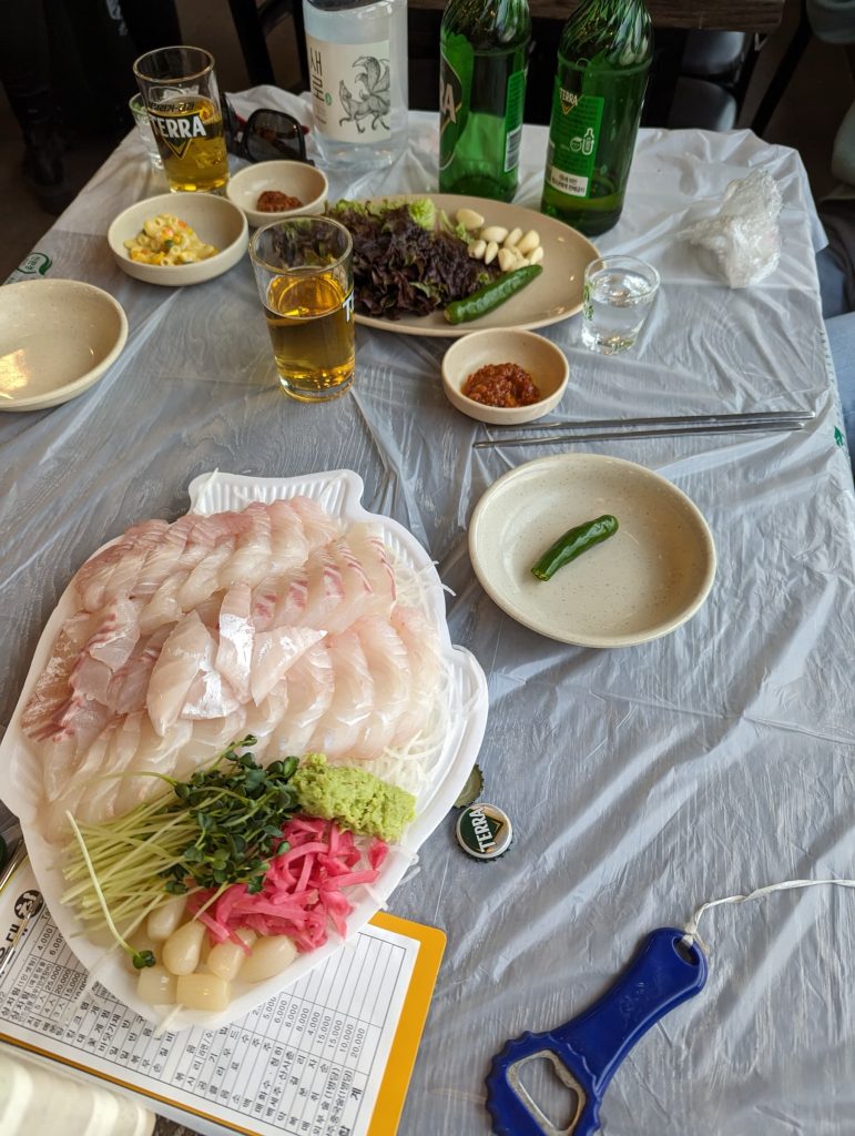 A table with food and beer. The main platter is full of a white fish, cut raw with condiments on the side— wasabi, pickled ginger, garlic, and a small green (I don't remember what this was).