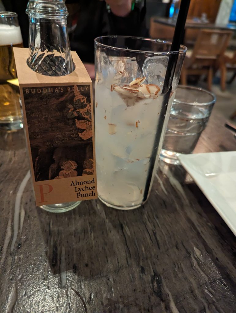 A drink in a tall glass sits next to a bottle with the label "Buddah's Belly Almond Lychee Punch". The drink is cloudy, and has shaved almonds.