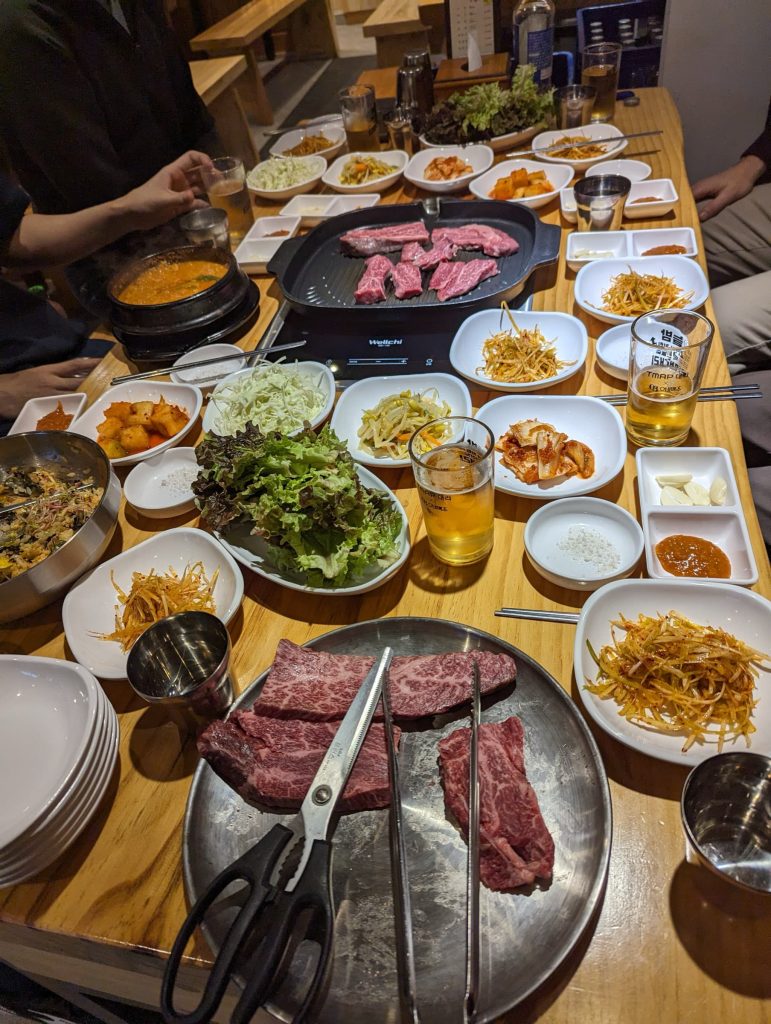 A restaurant table with grill in the middle, absolutely packed with small plates of side dishes and beef.