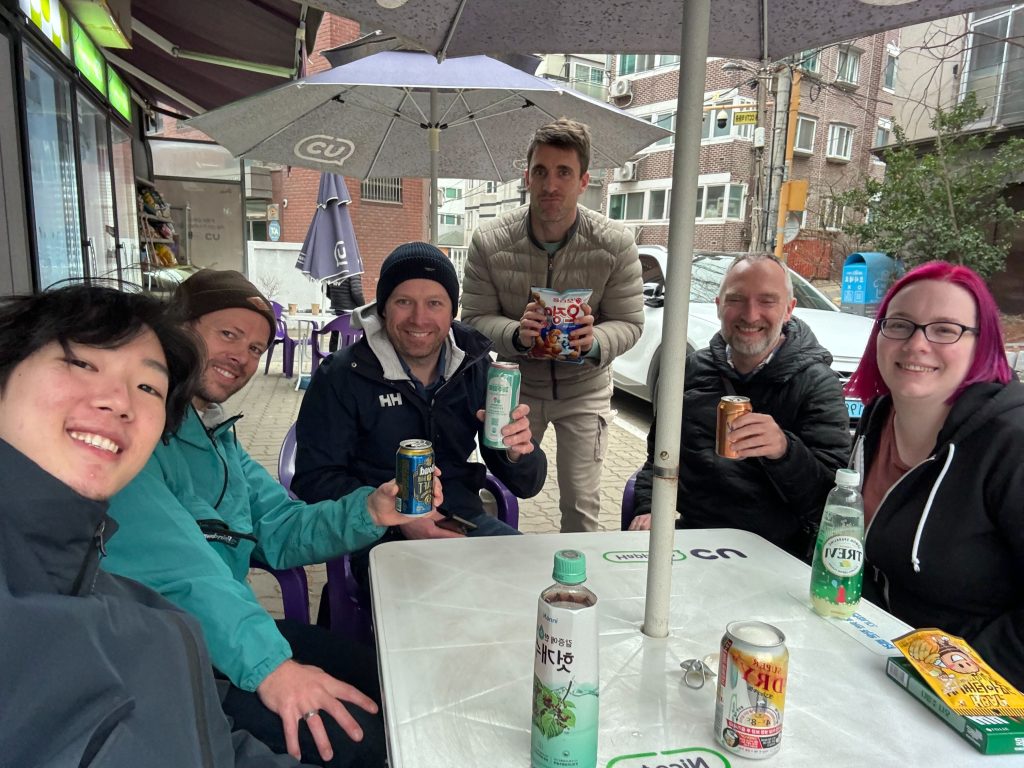 6 people sitting around a plastic table, some raising drinks to the camera.