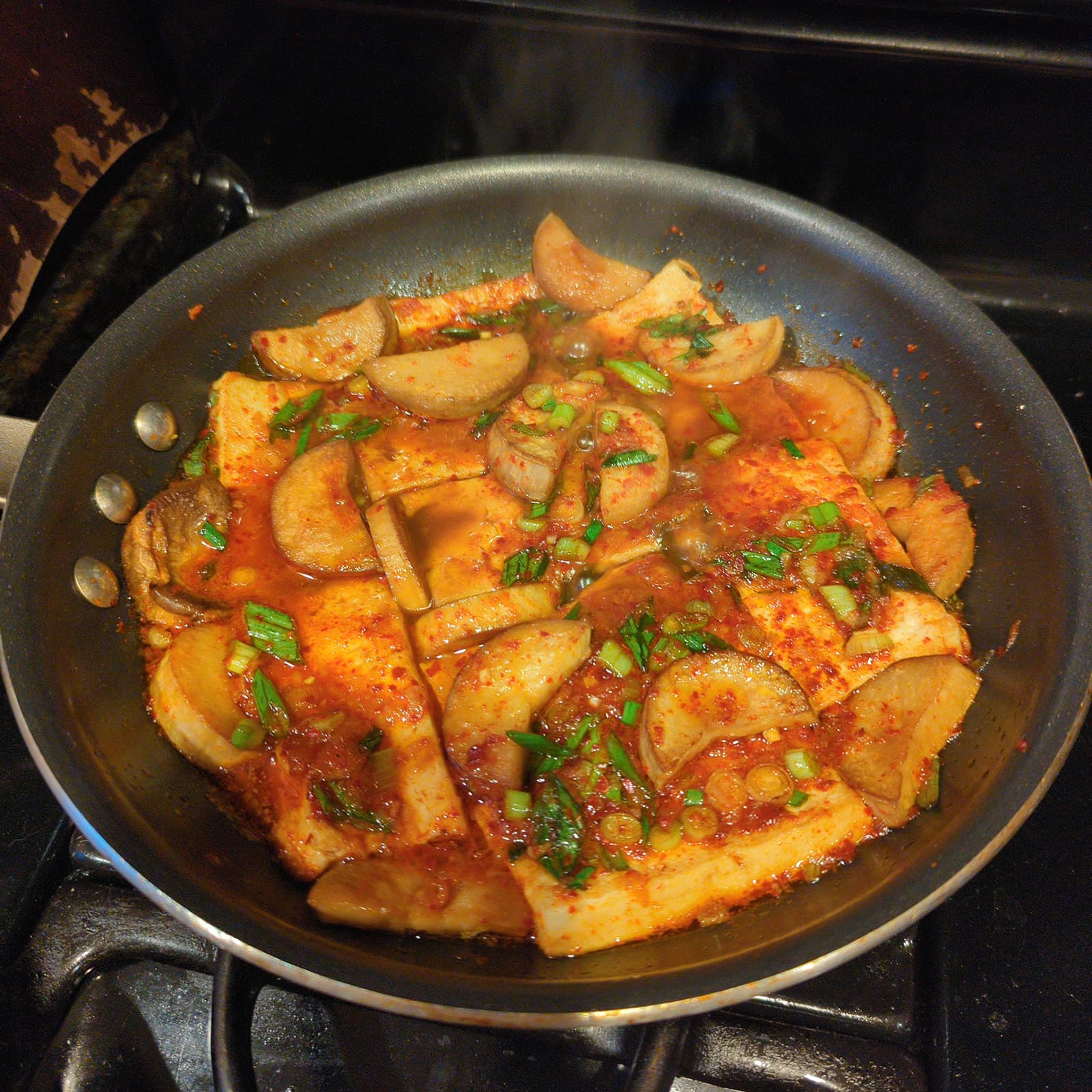 A frying pan on the stove with tofu and mushroom slices in a red sauce, with some steam coming off the food.