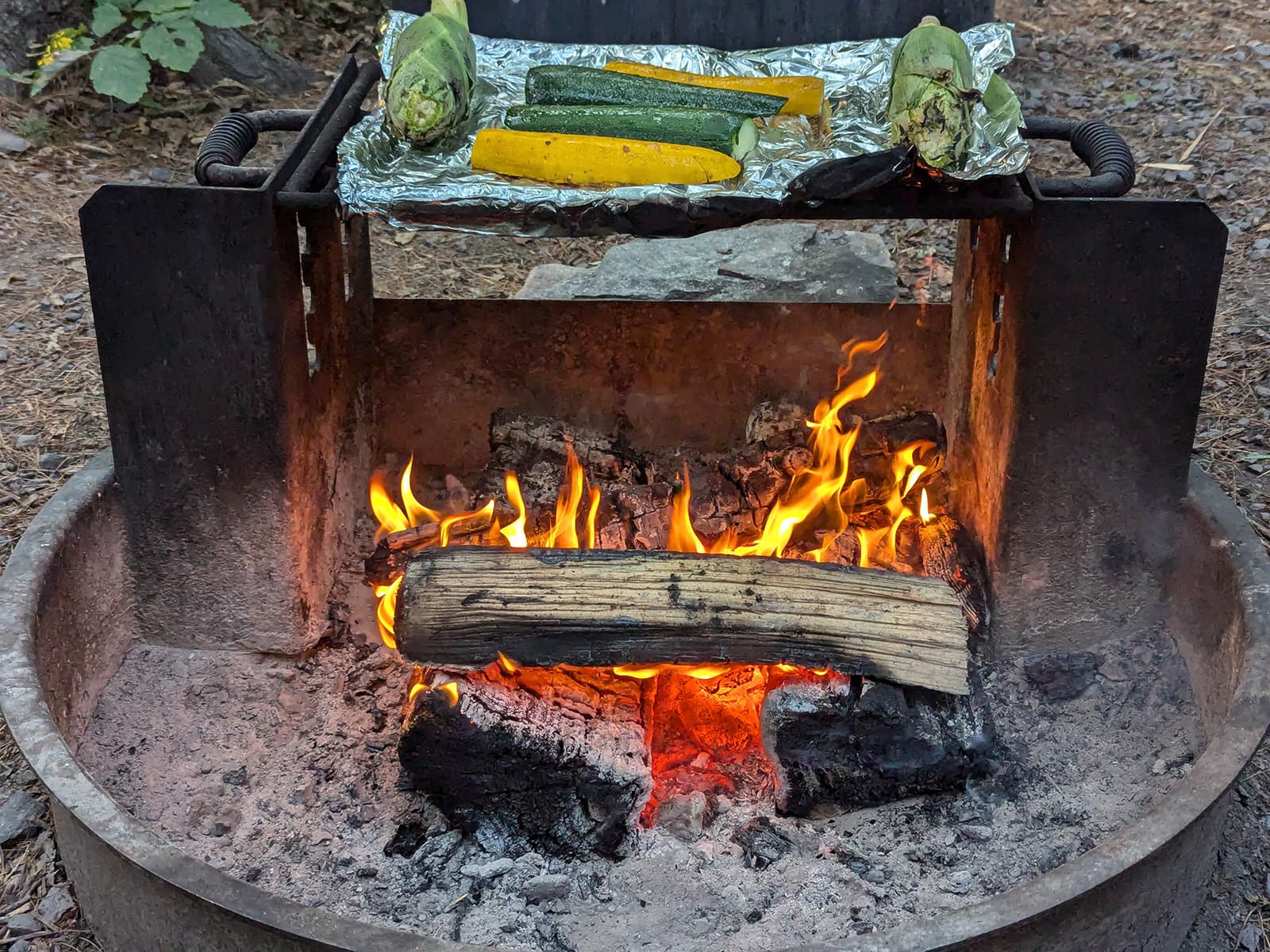 Fire in a fire pit, with zucchini and corn on the grill above.