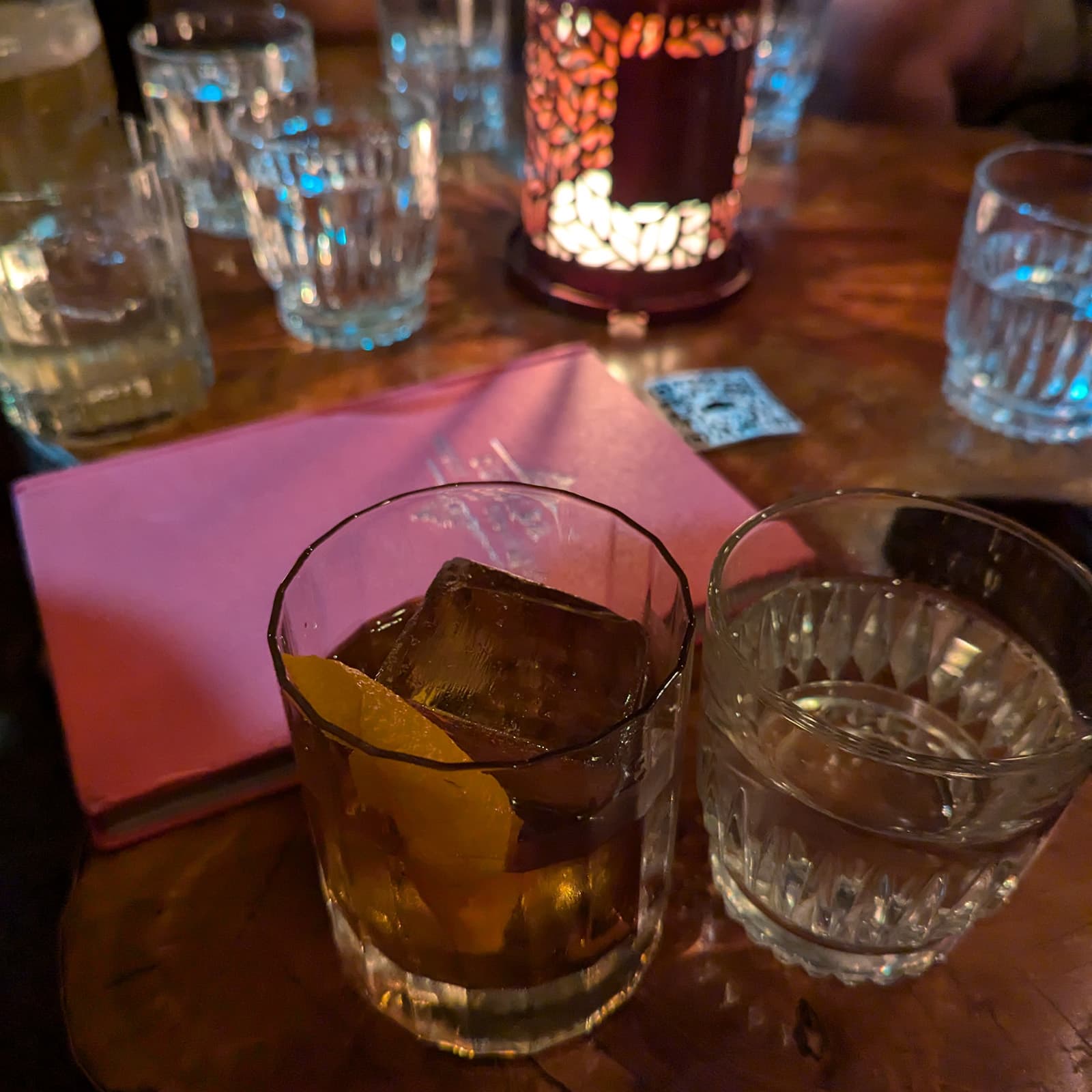 A candlelit table with multiple glasses, focus is on a short whiskey cocktail with a large ice cube and orange peel.
