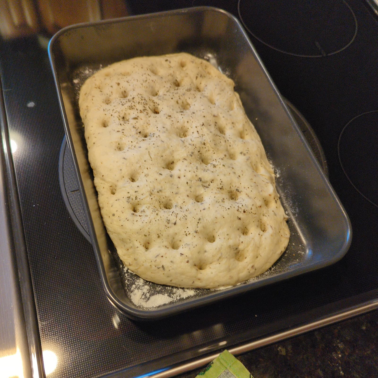 A baking dish with dough, spread out and poked with deep indents, covered in herbs.