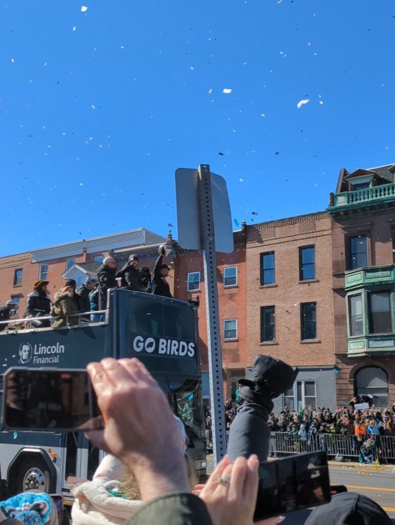 Street lined with crowds as a double-decker bus goes by, guy in the bus is raising a trophy & everyone is cheering.