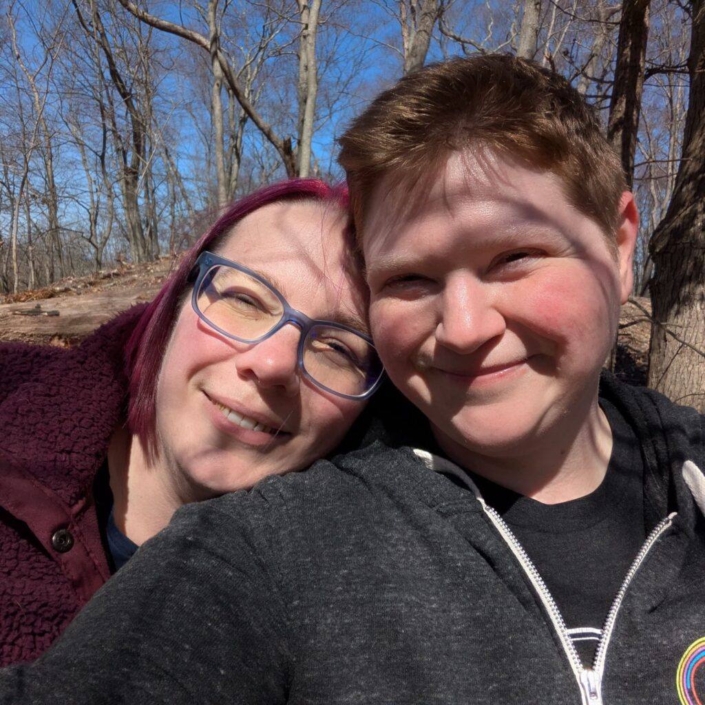 A selfie of Mel & I in a winter forest, just bare trees in the background. It's sunny, so we're smiling and squinting into the camera.