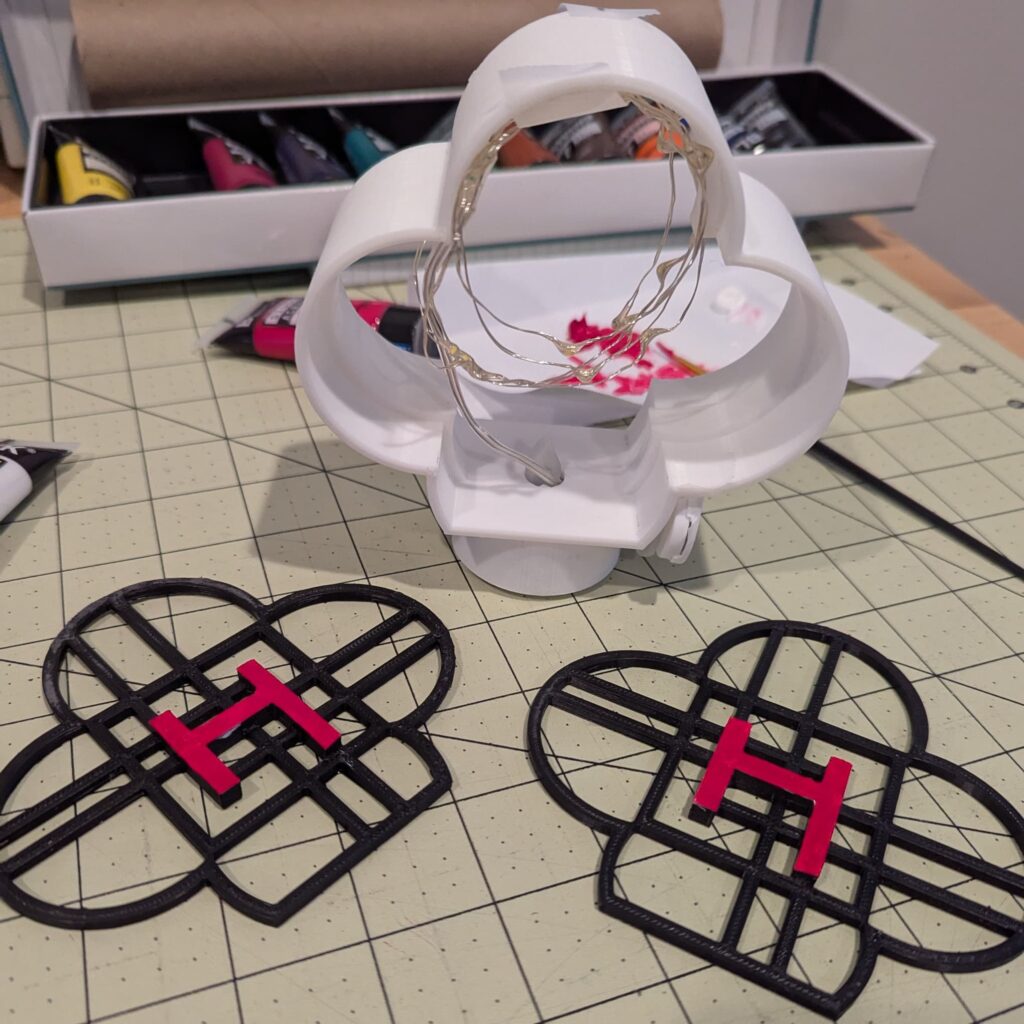 A craft workspace with a 3d print frame with lights inside, and 2 3d prints with the Huntrix symbol, painted pink. Paint and paintbrushes in the background.